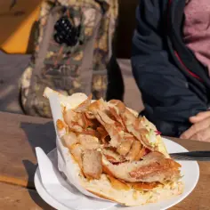 Pyszny kebab z soczystym mięsem, podany na talerzu, czeka na zjedzenie. Obok widać plecak i siedzącą osobę.