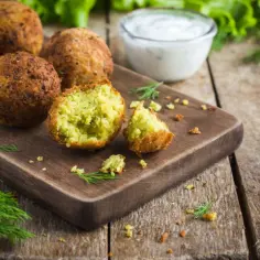 Falafel kebab co to? Chrupiące kulki falafela na desce, jedna przekrojona, z sosem i ziołami.