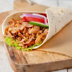 Pyszny kebab w piekarniku, zawinięty w tortillę z soczystym mięsem, sałatą, pomidorem, ogórkiem i czerwoną cebulą, podany na desce.