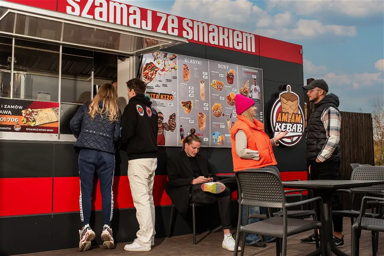 Ludzie stoją przy budce z kebabem, patrząc na menu. Zastanawiają się, ile można zarobić na kebabie, analizując ceny i popularność dań.
