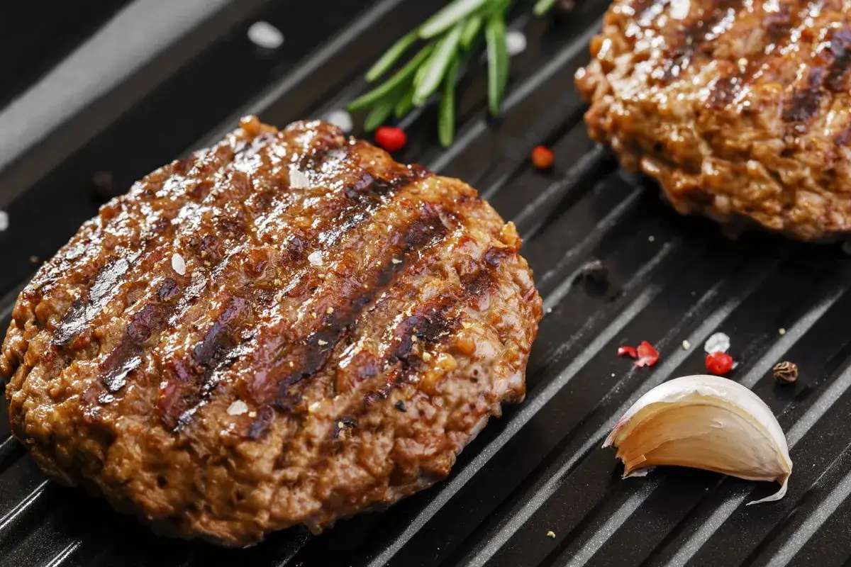 Soczyste kotlety wołowe na grillu, idealne na burgery. W tle gałązka rozmarynu i ząbek czosnku.