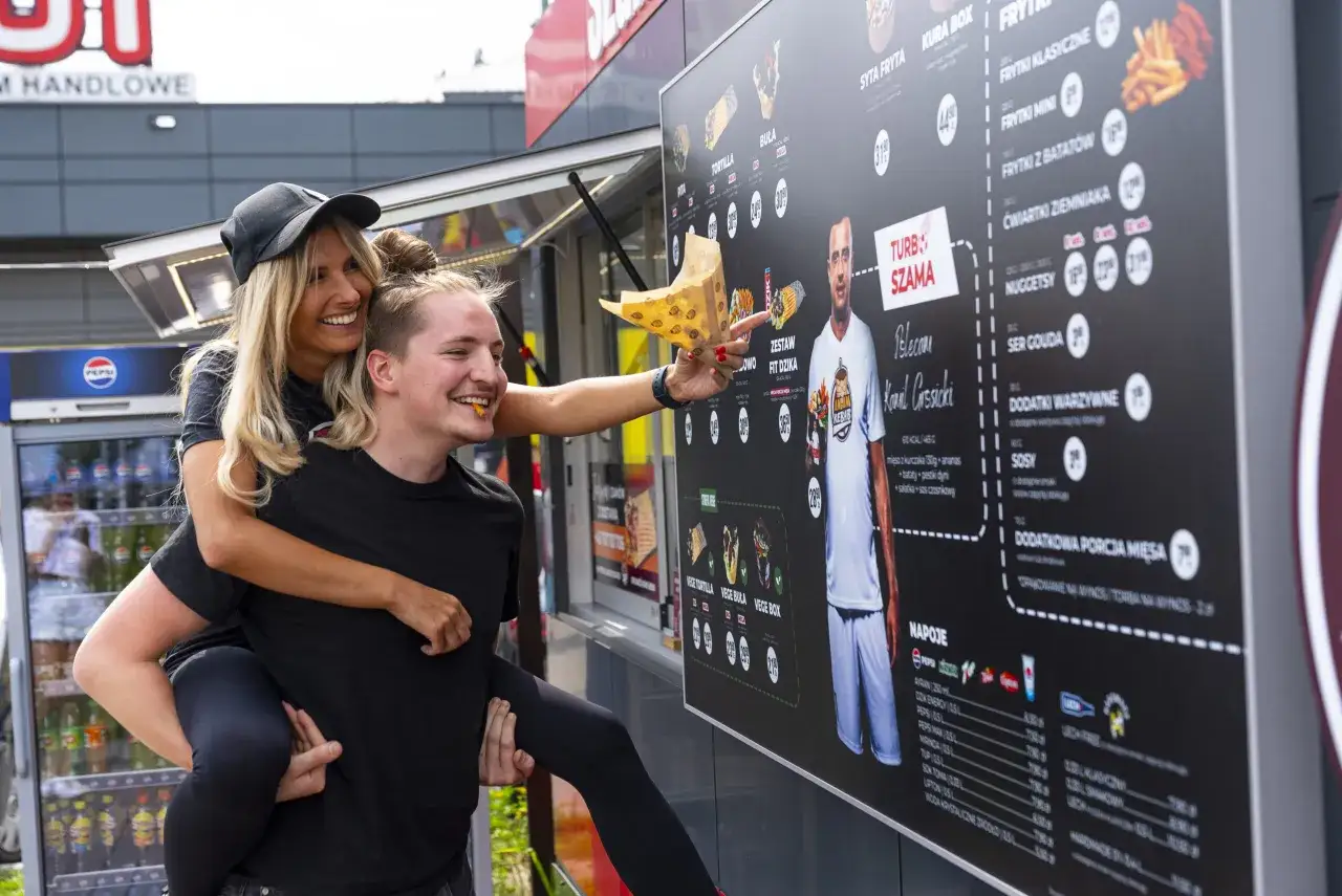 Para pozuje przy menu food trucka, zastanawiając się, ile można zarobić na kebabie. Ona niesie go na barana, on trzyma kawałek jedzenia.